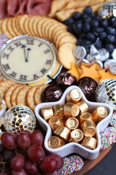 a platter filled with crackers, grapes, and other snacks next to a clock