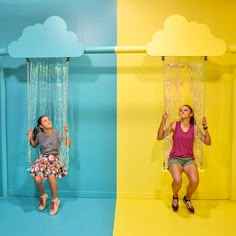 two women sitting on swings in front of a blue and yellow wall with clouds above them