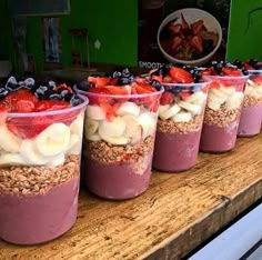 four cups filled with fruit and granola on top of a wooden table in front of a green wall