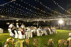 a group of people sitting at tables in the middle of a field with lights strung overhead