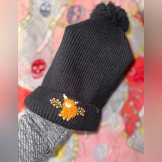 a person's hand wearing a black knitted hat with an embroidered deer on it