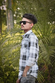 a young man wearing sunglasses standing in front of some plants and trees with his hands in his pockets