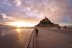 people are walking along the boardwalk near an old castle in the distance, at sunset