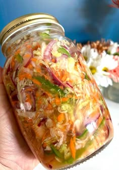 a hand holding a mason jar filled with vegetables and other food items on a table