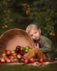 a young boy sitting on the ground next to a basket full of apples and holding an apple