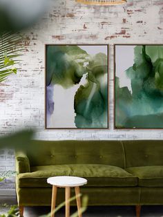 two paintings hanging on the wall above a green couch in a room with brick walls