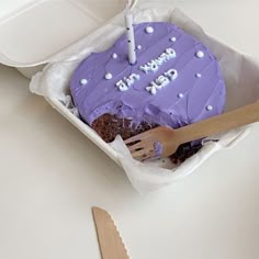 a purple birthday cake in a box with a knife and fork on the table next to it