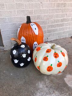 three painted pumpkins sitting on the ground next to a brick wall with ghost faces