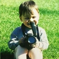 a little boy sitting in the grass holding a camera and looking at it's lens