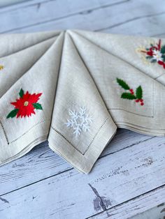 four napkins with embroidered holly and poinsettis on them sitting on a white wooden surface