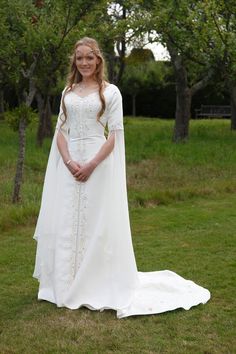 a woman wearing a white dress and cape standing in the grass with trees behind her