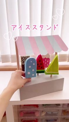 a person is holding up a small toy house in front of a window with pink and white striped awnings