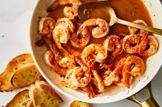 a bowl filled with shrimp and garlic bread next to a spoon on top of a table