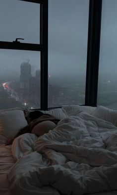 an unmade bed in front of a large window with city lights on the other side