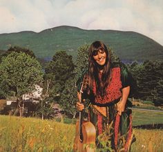 a woman standing in a field holding a guitar and smiling at the camera with mountains in the background