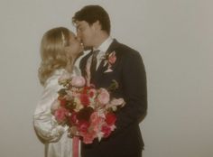 a bride and groom kissing each other in front of a white wall with flowers on it