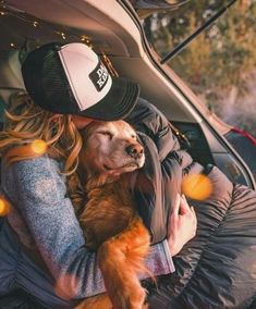 a woman sitting in the back seat of a car holding a brown dog and wearing a hat