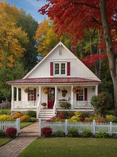 a white house with red shutters and flowers on the front yard, surrounded by trees