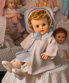 an old fashioned baby doll sitting on top of a bed next to other dolls and teddy bears