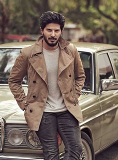 a man standing in front of a car wearing a trench coat and looking at the camera