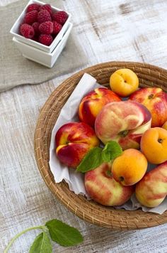 a basket filled with lots of ripe peaches on top of a table next to raspberries