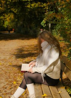 a woman is sitting on a bench reading a book in the fall leaves and looking at her phone