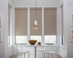 a dining room table with chairs and a bowl of fruit on it in front of two windows