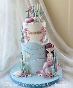 a three tiered cake decorated with mermaids and seashells on a blue plate