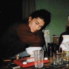 two people sitting at a table with cups and glasses on it, one person leaning over the table