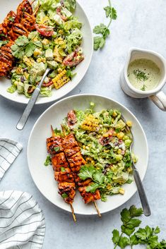 two plates filled with chicken skewers next to a bowl of salad and a cup of dressing