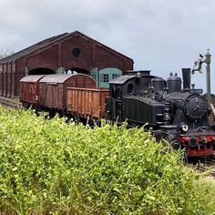 an old fashioned train is parked in front of a brick building and shrubbery on the tracks