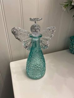 a glass bottle with an angel decoration on the top sitting on a white table next to a potted plant