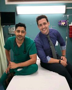 two men in scrubs are sitting on a hospital bed smiling at the camera,