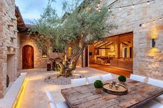 an outdoor dining area with table, chairs and tree in front of stone building at dusk