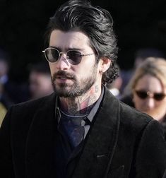 a man with black hair wearing sunglasses and a suit jacket walking in front of other people