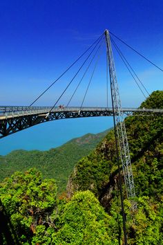 a bridge over water surrounded by lush green trees and mountains under a blue cloudless sky