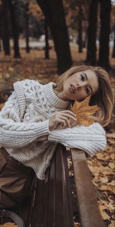 a woman sitting on top of a wooden bench next to a forest filled with leaves