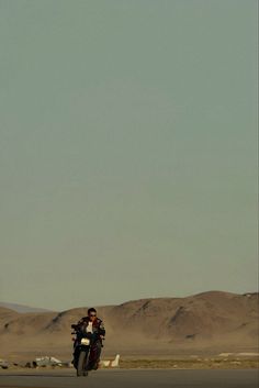 a man riding a motorcycle down the middle of a desert road with mountains in the background