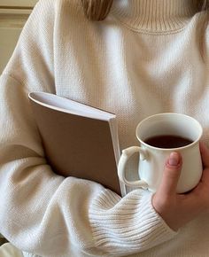 a woman is holding a book and a cup of coffee in her hands while wearing a white sweater