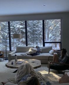 two people sitting on couches in a living room with snow covered trees outside the window