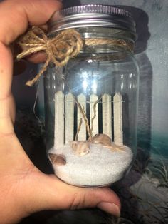 a hand holding a glass jar filled with sand and sea shells on top of it