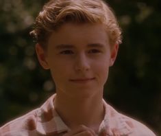 a young man with blonde hair wearing a pink and white shirt smiling at the camera