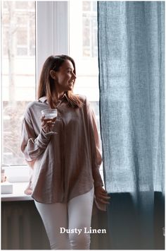 a woman standing in front of a window holding a wine glass and looking out the window