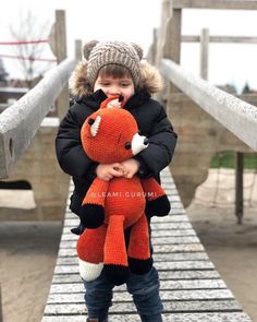 a little boy holding a stuffed animal on top of a wooden bridge in the snow