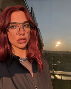 a woman with red hair and glasses standing in front of a window looking at the camera