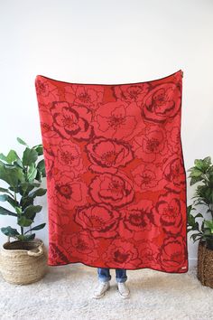 a woman is holding up a red floral print blanket in front of some potted plants