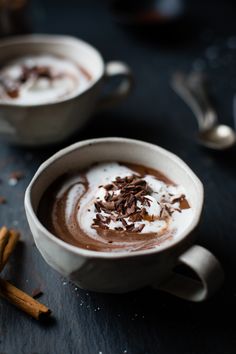 two cups of hot chocolate with whipped cream and cinnamon sticks on the table next to them