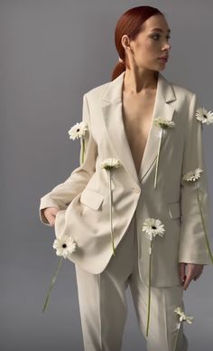 a woman with red hair wearing a white suit and flowers on her lapel, standing in front of a gray background