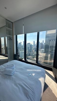 a bed with white sheets and pillows in a bedroom next to large windows overlooking the city