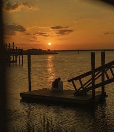 two people sitting on a dock in the middle of water at sunset, with sun setting behind them